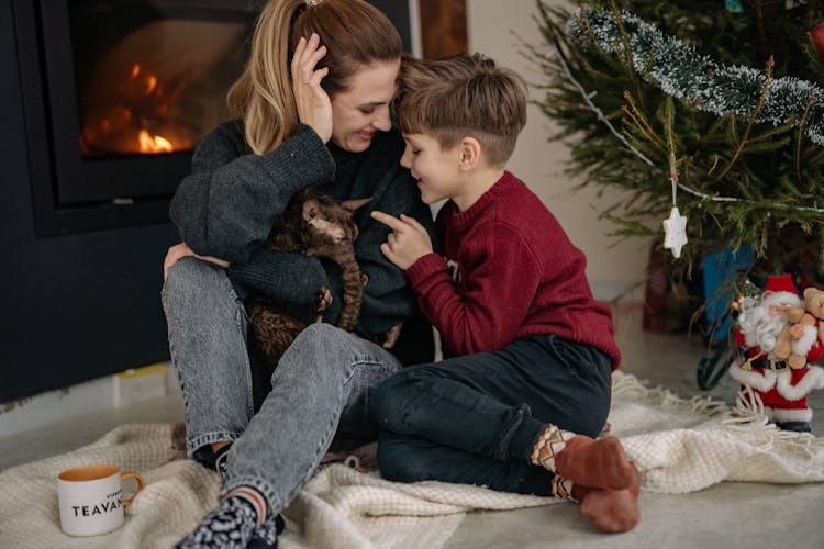 Mother Holding A Cat With Her Son Sitting By The Fireplace At Christmas 