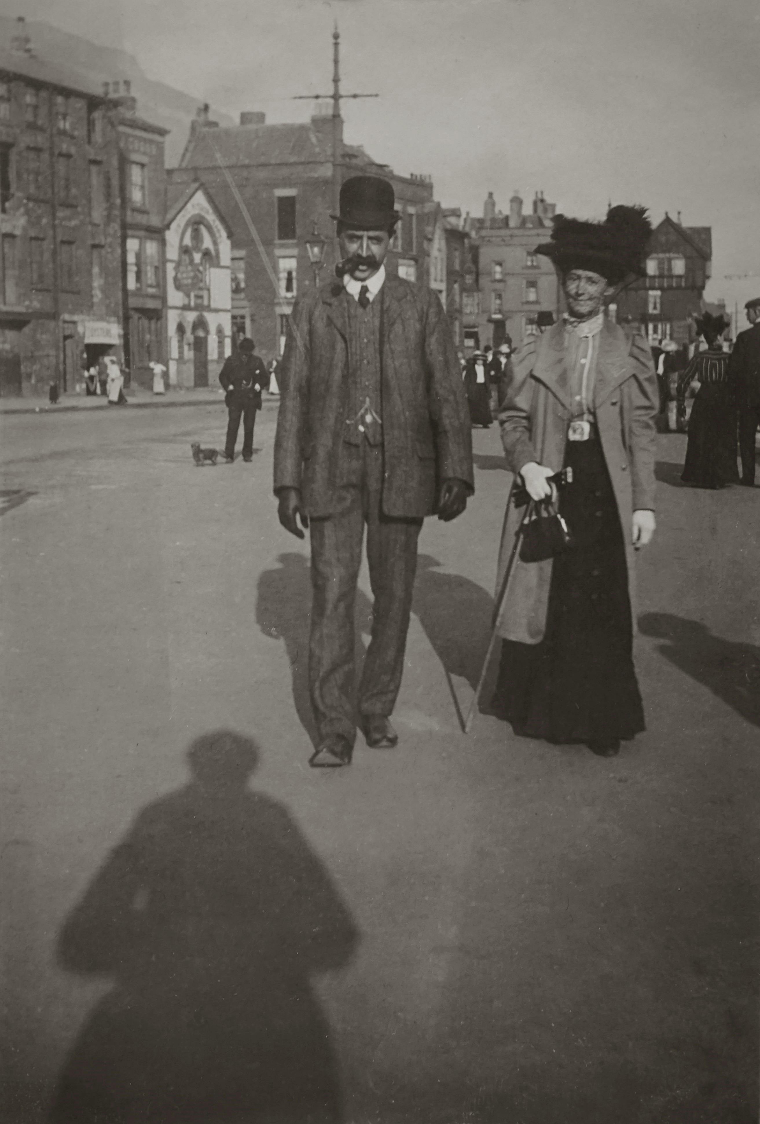 Elegant couple in vintage attire walking on a historic street, early 20th century.