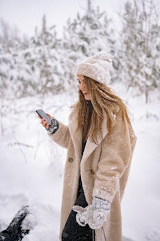 Smiling woman in winter coat and hat using mobile phone in snowy forest.