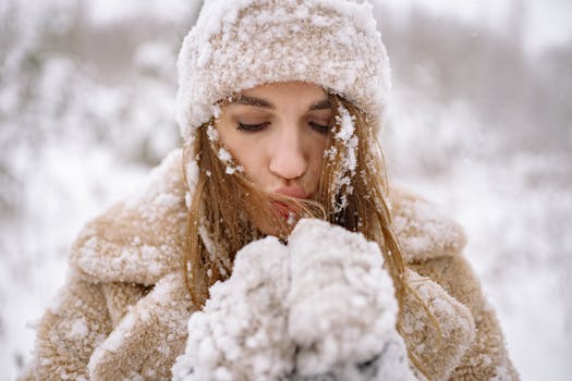Close-up of a woman in winter attire surrounded by snow, blowing warm air into her hands.