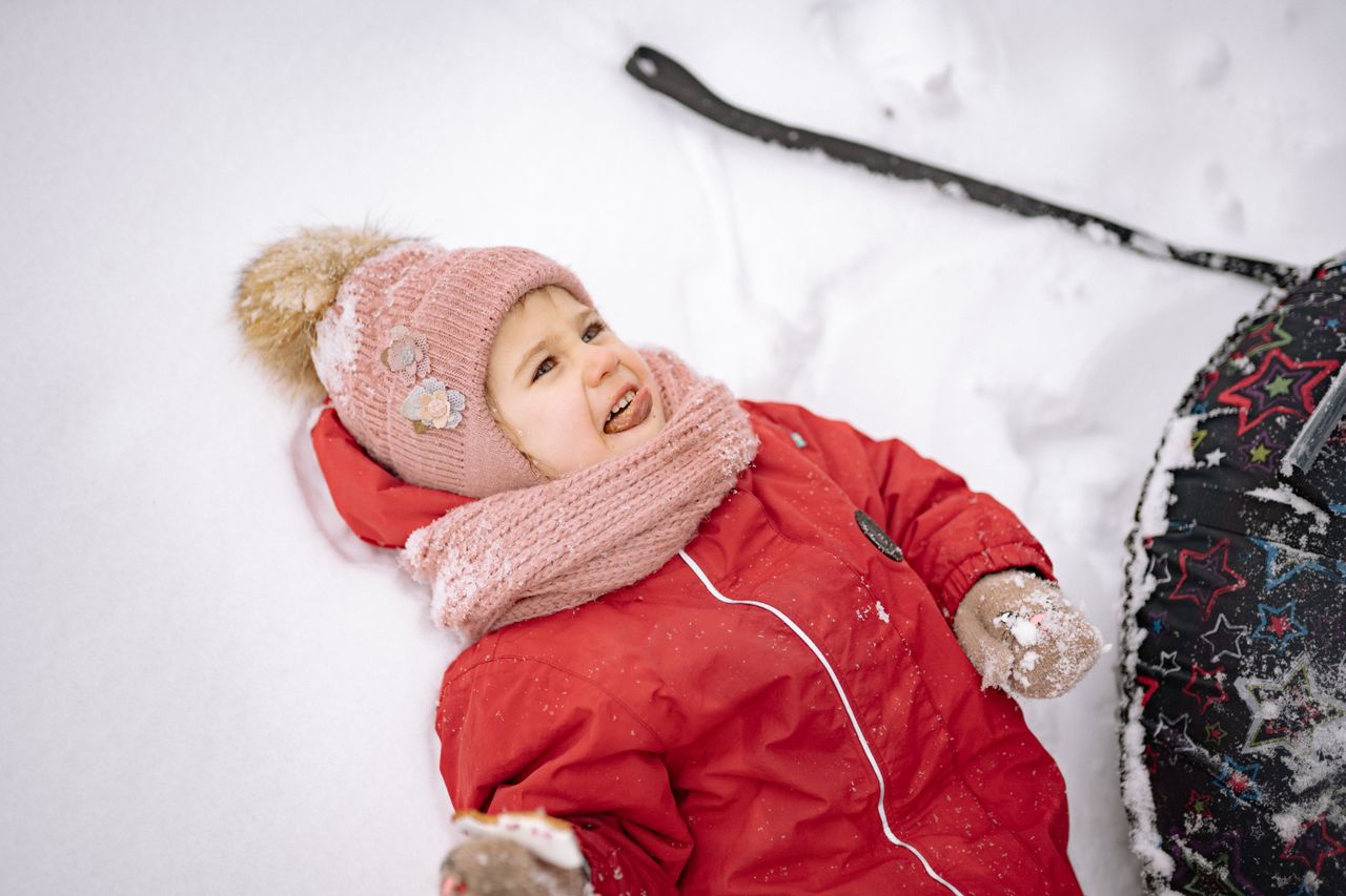 Smiling girl in a red snowsuit, pink knit hat and scarf lying in fresh snow
