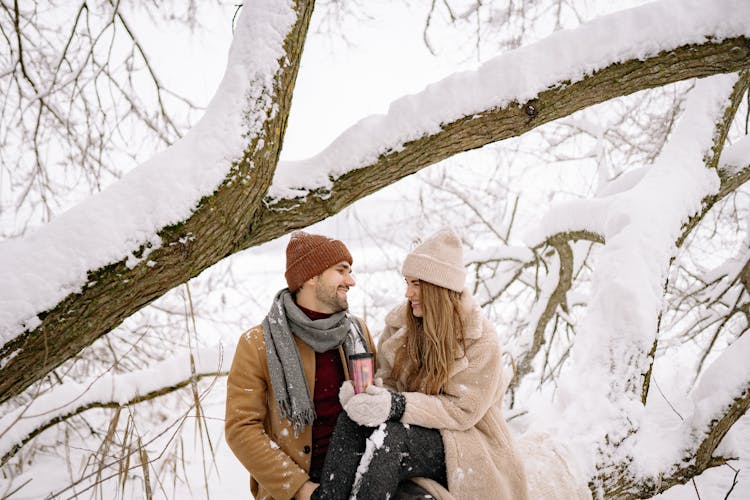 A Lovely Couple In Winter Clothes Looking At Each Other Under The Snow-Covered Tree Branch