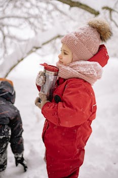 A child warmly dressed in red clothing holds a drink while enjoying the snowy winter outdoors.