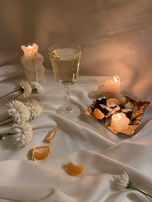 Free White flowers and slices of oranges placed on white crumpled textile with wineglass of champagne near tray with candies in stylish room with burning candles Stock Photo