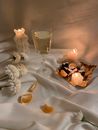 White flowers and slices of oranges placed on white crumpled textile with wineglass of champagne near tray with candies in stylish room with burning candles