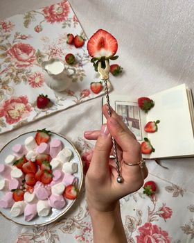 From above of unrecognizable female with strawberry on fork standing near plate full of marshmallows and jug of milk in light room with book