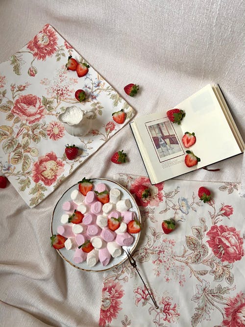 Top view of plate full of colorful marshmallows and slices of fresh strawberry placed on textile with jug of milk and book in stylish room