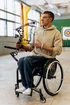 A male archer in a wheelchair practicing archery indoors, aiming his bow at a target.