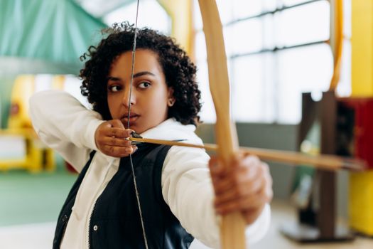 Young woman with a determined expression practicing archery in an indoor setting.