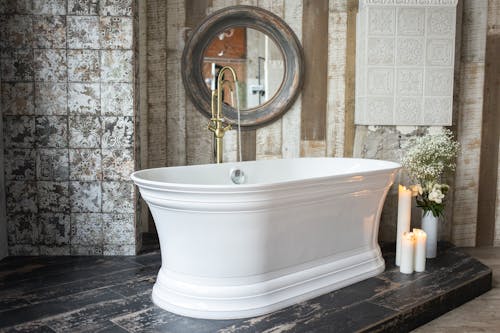 White bath with golden faucet placed near burning candles and green plants in vase near wall with mirror in modern bathroom