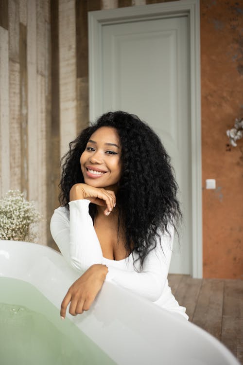 Positive African American female smiling and looking at camera while sitting near tub with clear water during daily routine in bathroom