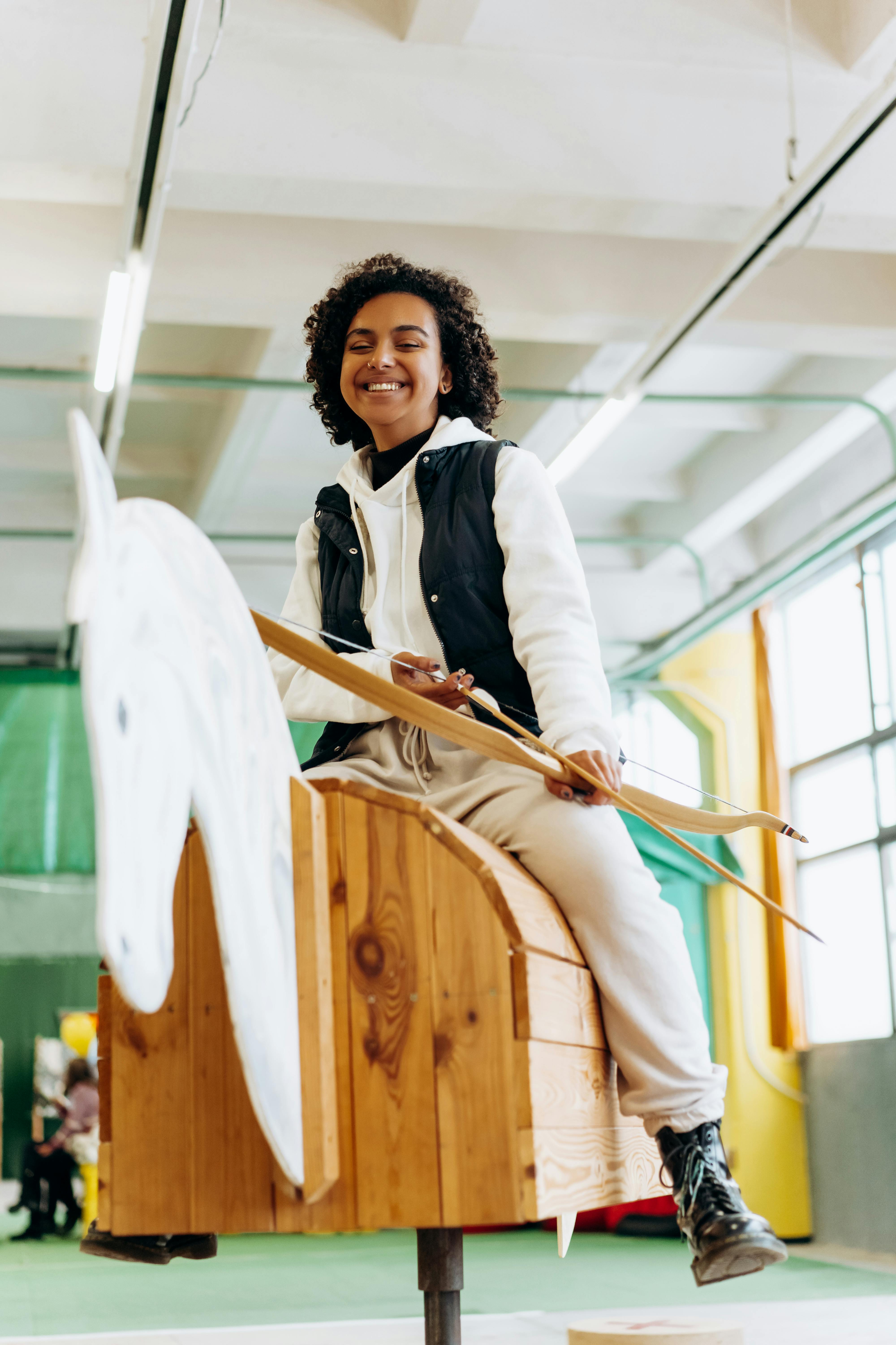 Happy young woman practicing archery indoors on a wooden horse simulator, smiling playfully.