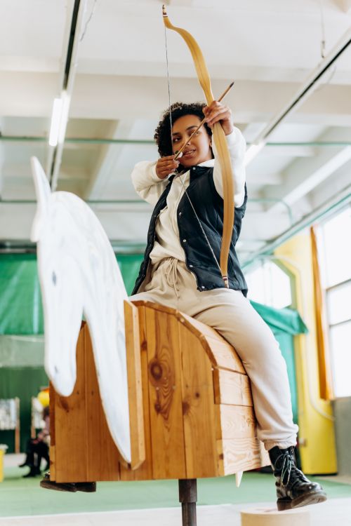 Free A young woman practicing archery indoors on a wooden horse, aiming confidently. Stock Photo