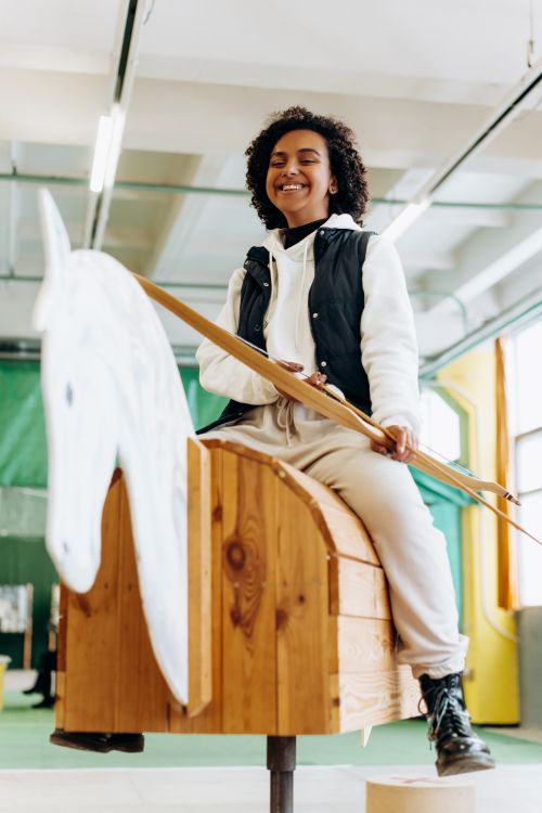 Free A smiling young woman practicing archery indoors on a wooden horse, embodying leisure and focus. Stock Photo