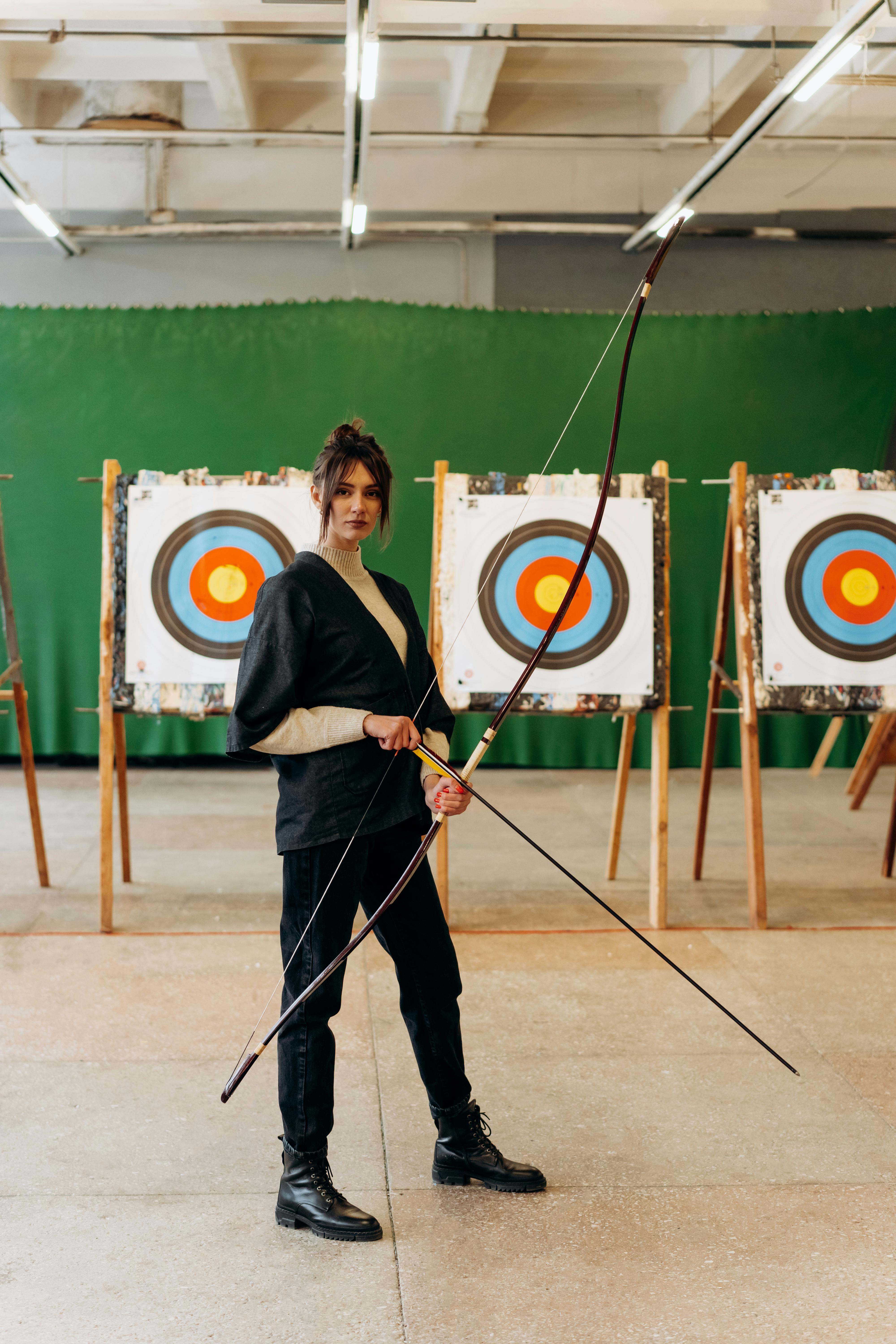 A young female archer standing confidently with a bow indoors, in front of colorful targets.