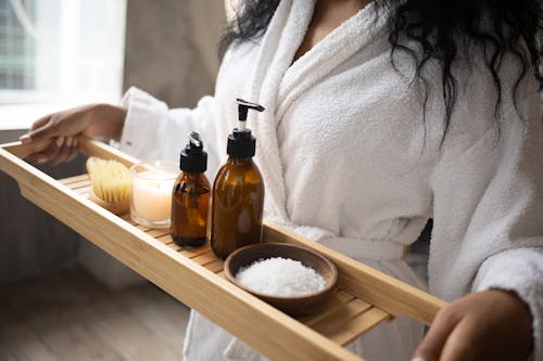 Unrecognizable African American female in white bathrobe with bottles of cosmetic products salt and burning candle on wooden tray in hands