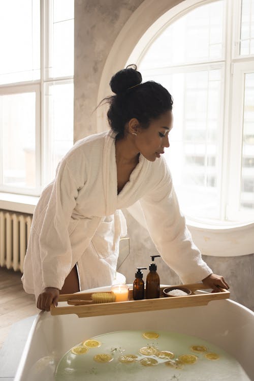 African American female in white bathrobe putting wooden tray with spa products on bathtub with lemon slices during skin care routine