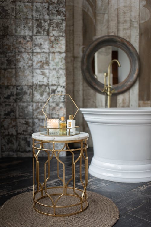 Glass box with spa supplies placed on small round table near white bathtub in stylish bathroom near wall with mirror