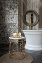 Glass box with spa supplies placed on small round table near white bathtub in stylish bathroom near wall with mirror