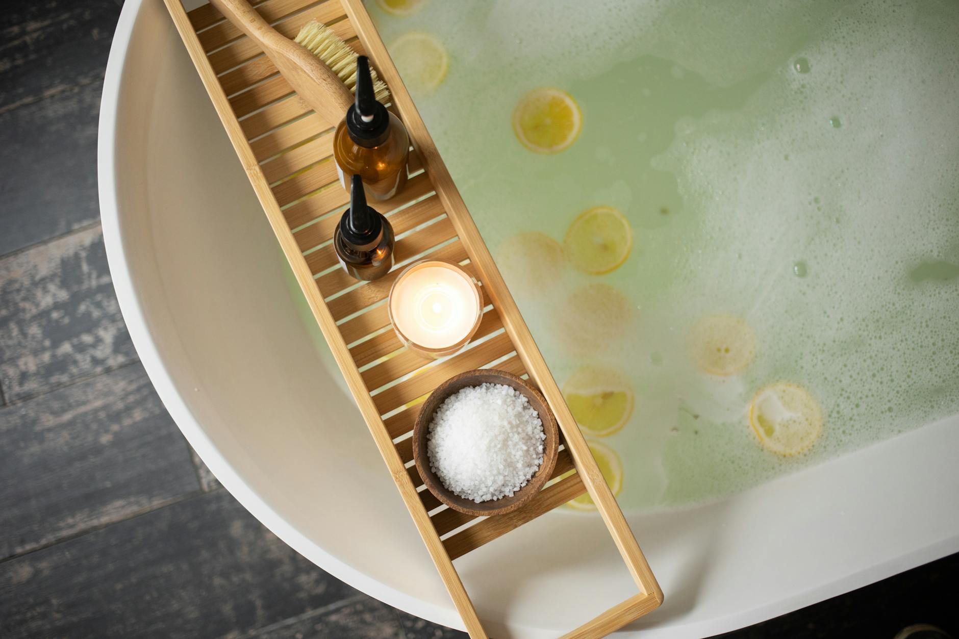 Top view of wooden tray with salt and bottles of cosmetic products placed on white tub with lemon slices on water surface