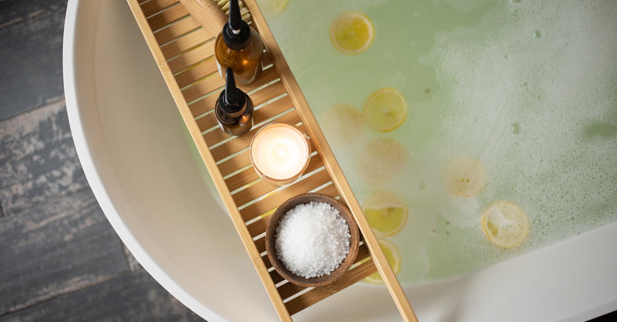 Top view of wooden tray with salt and bottles of cosmetic products placed on white tub with lemon slices on water surface