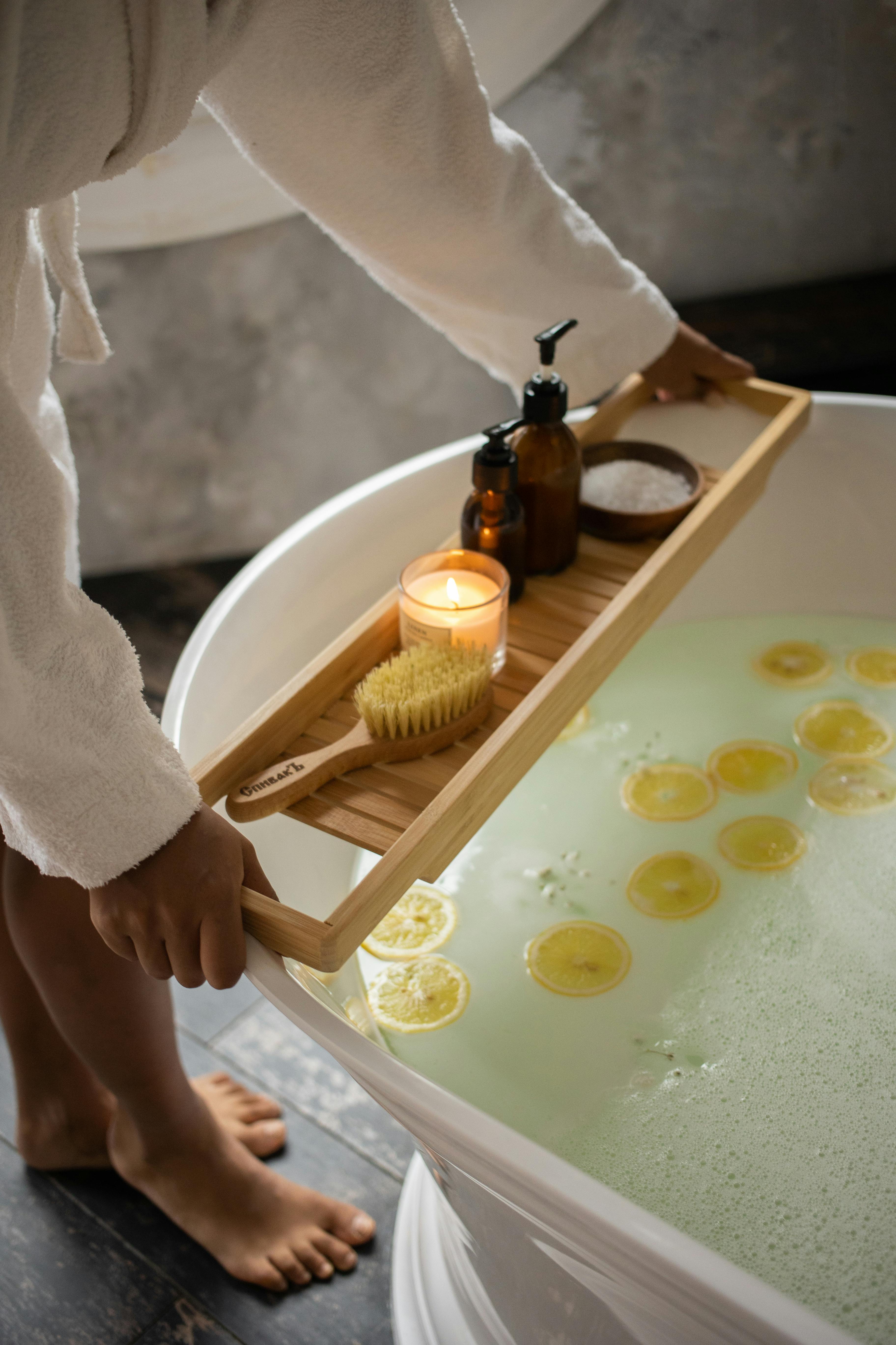 Free Unrecognizable barefoot female in bathrobe putting wooden tray with spa supplies on tub with lemon slices on water surface during skincare routine Stock Photo