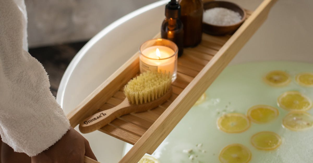 Unrecognizable barefoot female in bathrobe putting wooden tray with spa supplies on tub with lemon slices on water surface during skincare routine