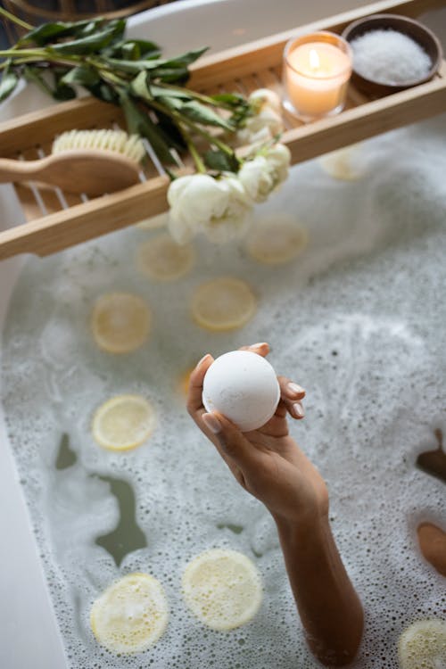 Free From above of unrecognizable female sitting in bath with lemon slices on water surface near wooden tray with salt and burning candle decorated with flowers during daily routine Stock Photo