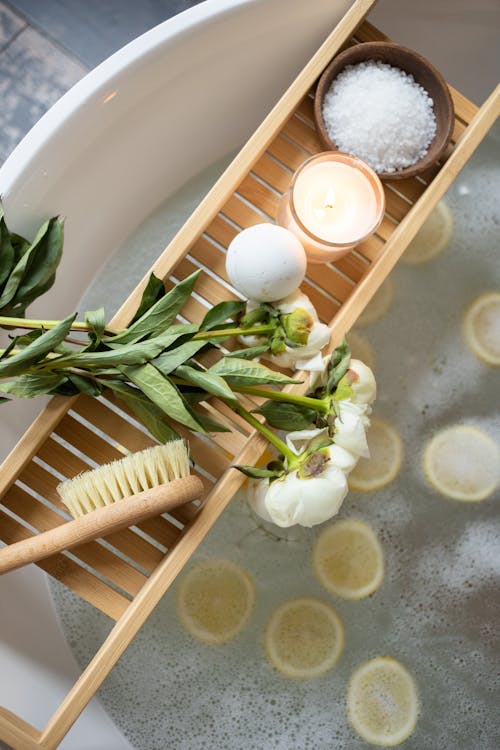 Top view of wooden tray with bath bomb and burning candle placed near salt and flowers on tub with lemon slices on water