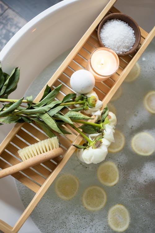 Free Top view of wooden tray with bath bomb and burning candle placed near salt and flowers on tub with lemon slices on water Stock Photo