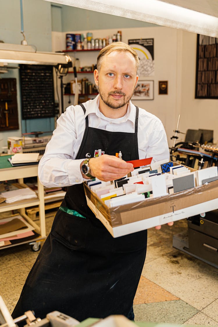 A Man In White Long Sleeves And Black Apron Holding A Box With Cards