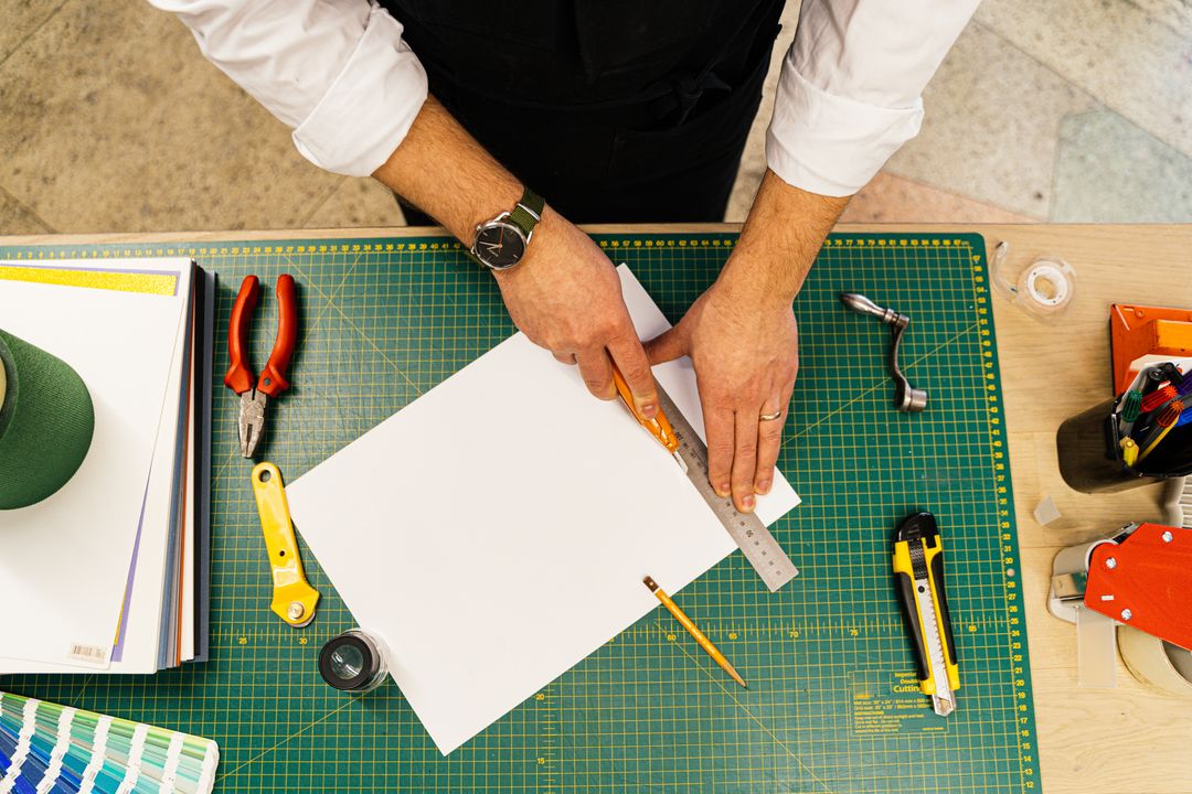 Top view of hands using a ruler and cutter on a work desk with various craft tools and paper.