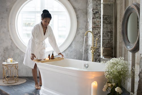 Full body of barefoot African America female in bathrobe putting wooden tray with spa supplies on bath during spa procedure