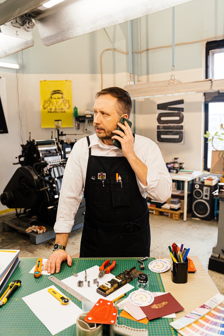 Graphic Designer Standing By A Desk In A Studio, Using Phone