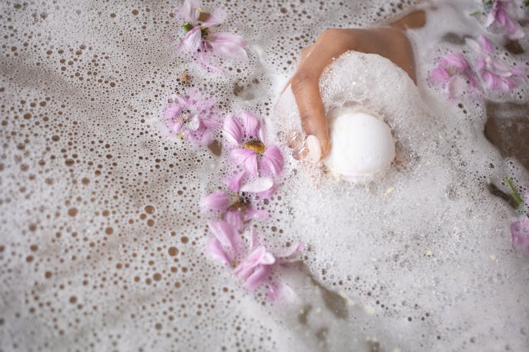 Woman Holding Ball Of Bath Salt In Water With Foam And Flowers