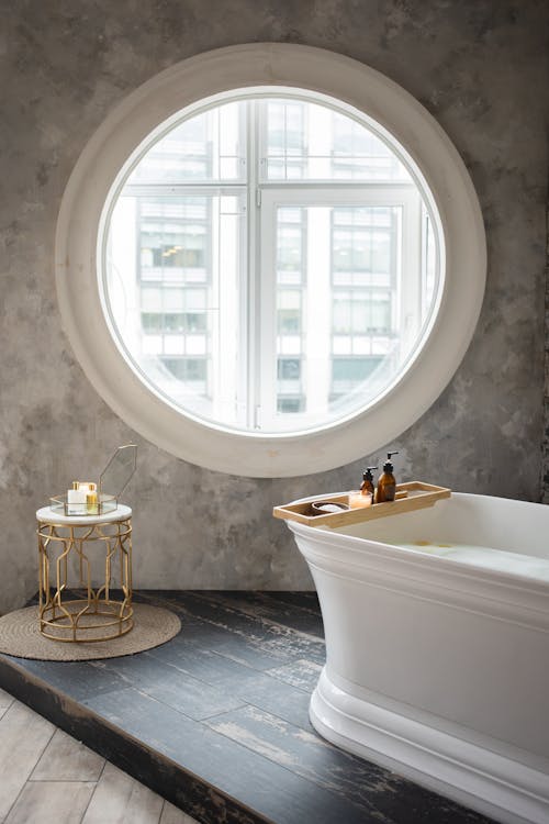 Interior of contemporary bathroom with white bathtub and small table placed against concrete wall with round window in daylight