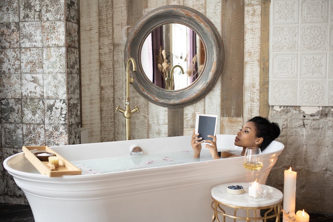 Free Young African American female reading book while taking bath in light bathroom with burning aromatic candles placed near round table with wineglass and berries on background of shabby stone walls Stock Photo