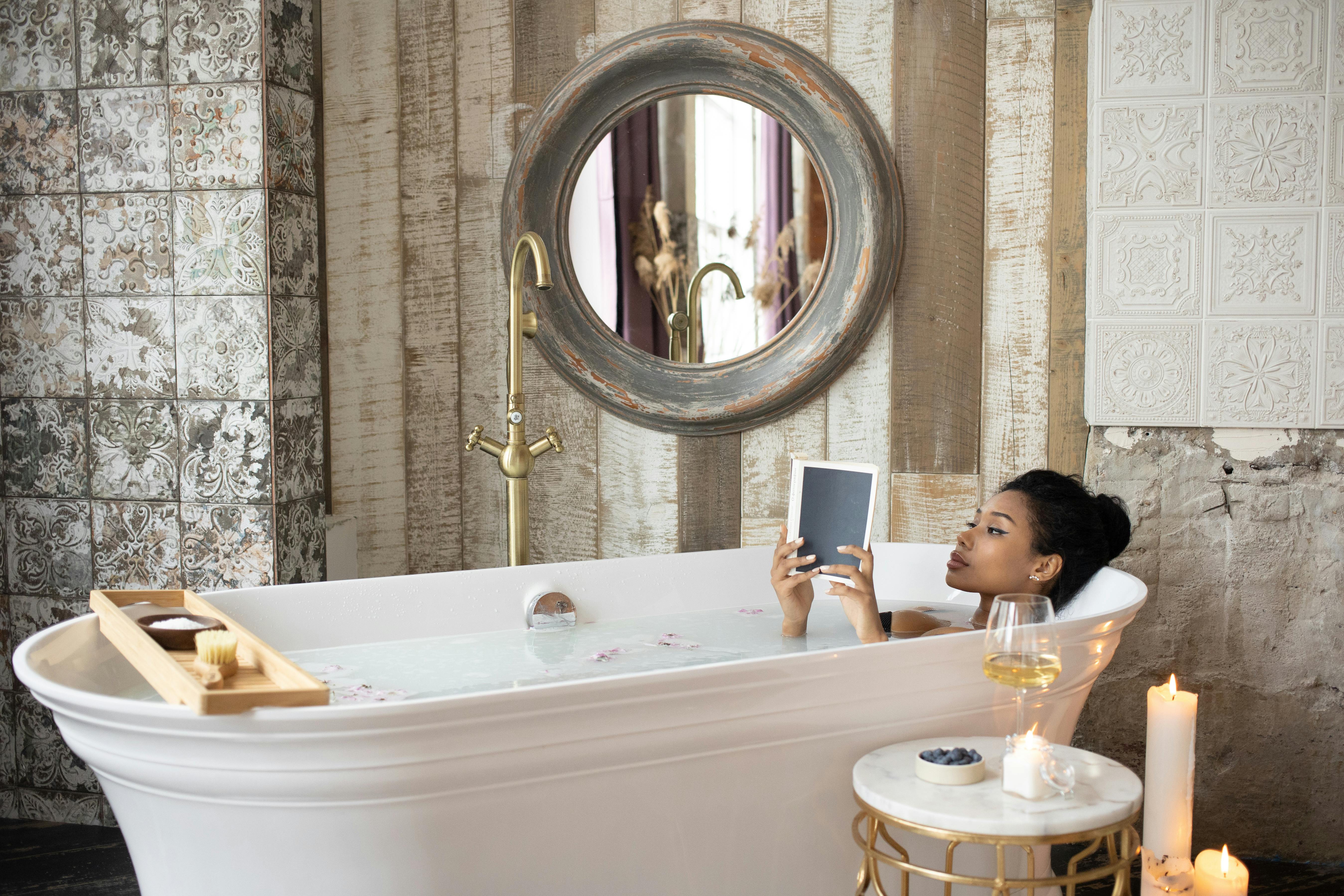 Young African American female reading book while taking bath in light bathroom with burning aromatic candles placed near round table with wineglass and berries on background of shabby stone walls