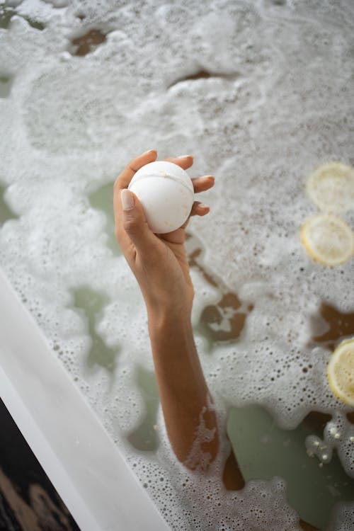 From above anonymous female holding ball of bath salt while taking bath with foam and slices of lemon
