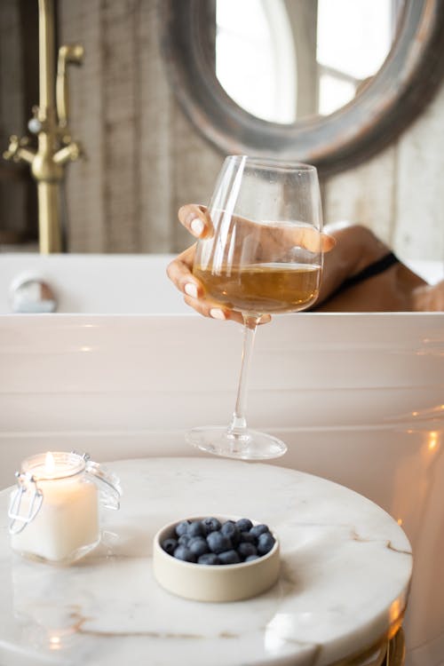 Free Unrecognizable female taking glass of champagne from round table with burning candle and bowl of berries while taking bath on blurred background against mirror Stock Photo