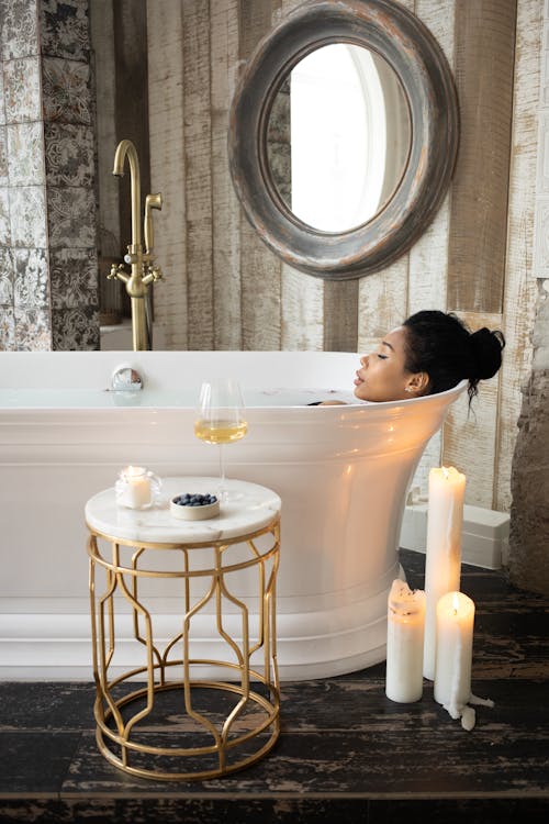 Side view of young ethnic female lying in bathtub with closed eyes in light bathroom with candles and table on background of mirror