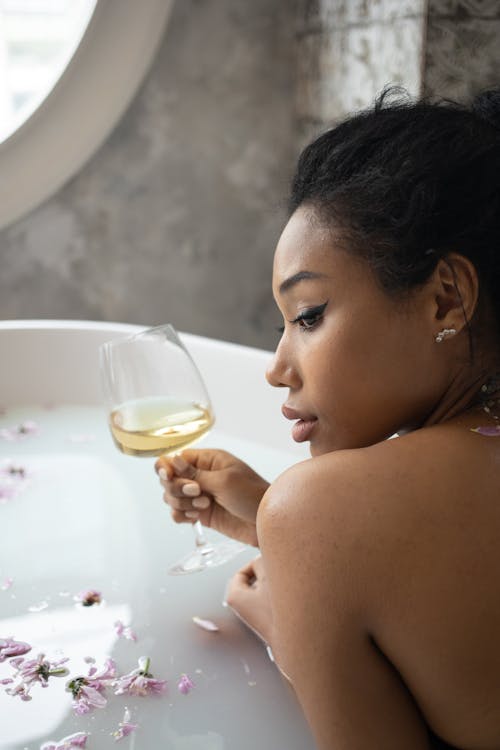 Young Black female with bare shoulder sipping wine form glass while sitting in bath with flowers against shabby walls with window