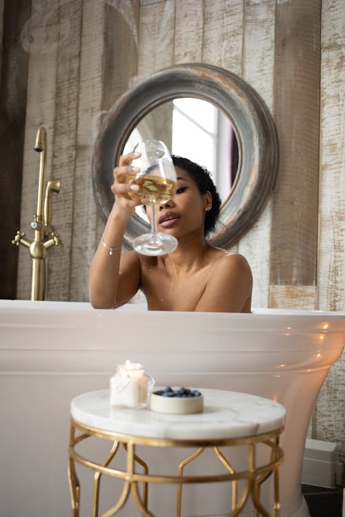 Free Young sensual ethnic female toasting with glass of white wine sitting in bathtub in light bathroom with candles and table on background of round mirror Stock Photo