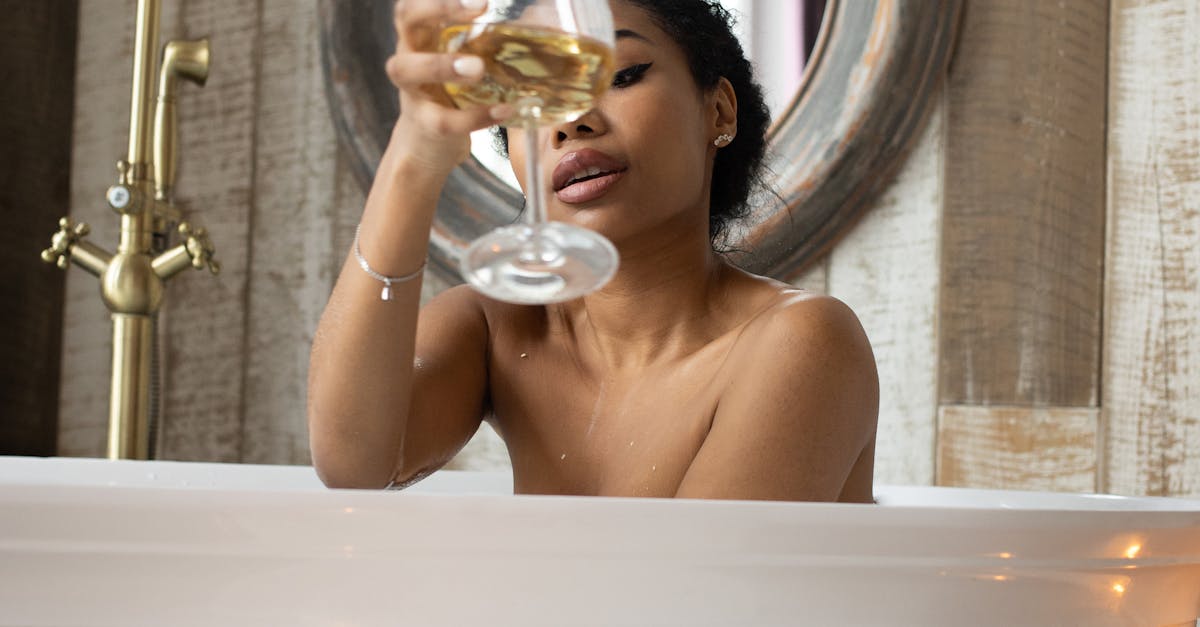 Young sensual ethnic female toasting with glass of white wine sitting in bathtub in light bathroom with candles and table on background of round mirror