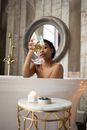 Young sensual ethnic female toasting with glass of white wine sitting in bathtub in light bathroom with candles and table on background of round mirror