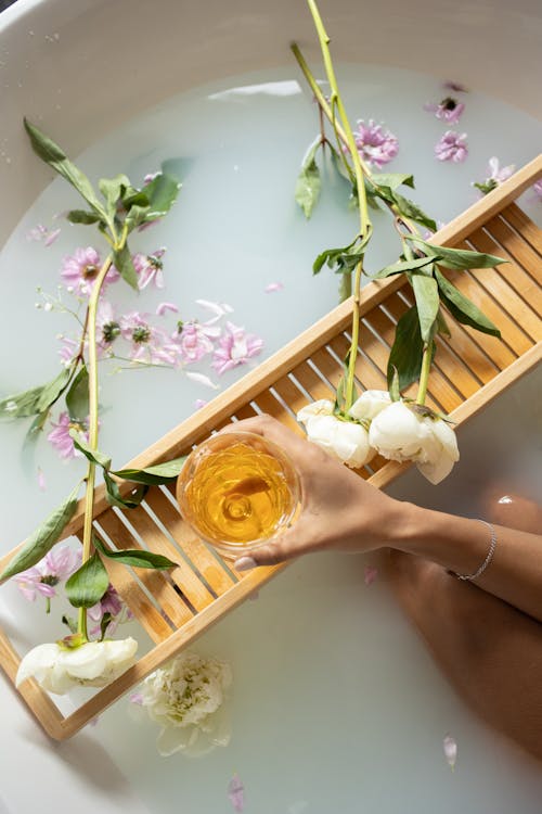 Top view of anonymous female with white wine sitting in bathtub with pink and white flowers