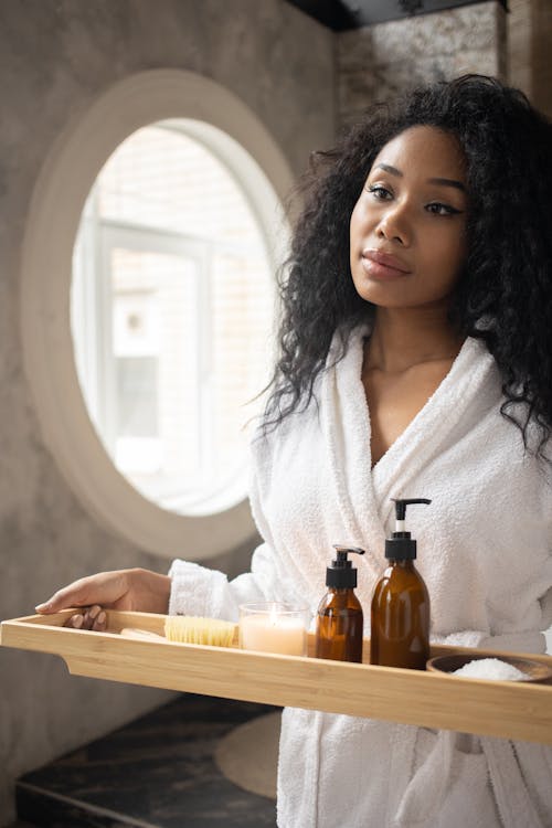 Free Young ethnic female in white bathrobe holding wooden tray with skin care products standing in bathroom and looking away Stock Photo