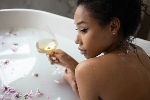 From above of young ethnic female holding glass of white wine while lounging in white water with petals in tub