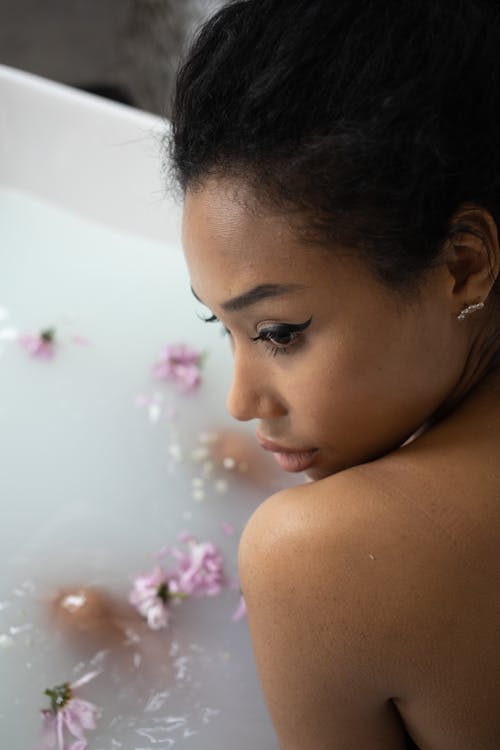 Charming ethnic female with makeup looking away while sitting in bathtub full of water with delicate flowers and petals on blurred background
