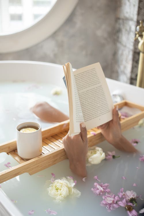 Free From above of unrecognizable female lying in bathtub full of water with delicate ivory and pink flowers and reading book on blurred background Stock Photo